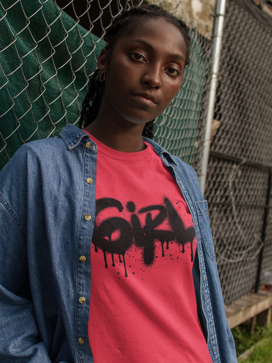 Una giovane donna con le treccine e un piercing al naso indossa una camicia di jeans azzurra aperta su una t-shirt rosso corallo. La maglietta presenta una stampa grafica nera in stile graffito che recita "GIRL" con un effetto gocciolante. Posa appoggiata a una recinzione metallica, con uno sfondo urbano sfocato.