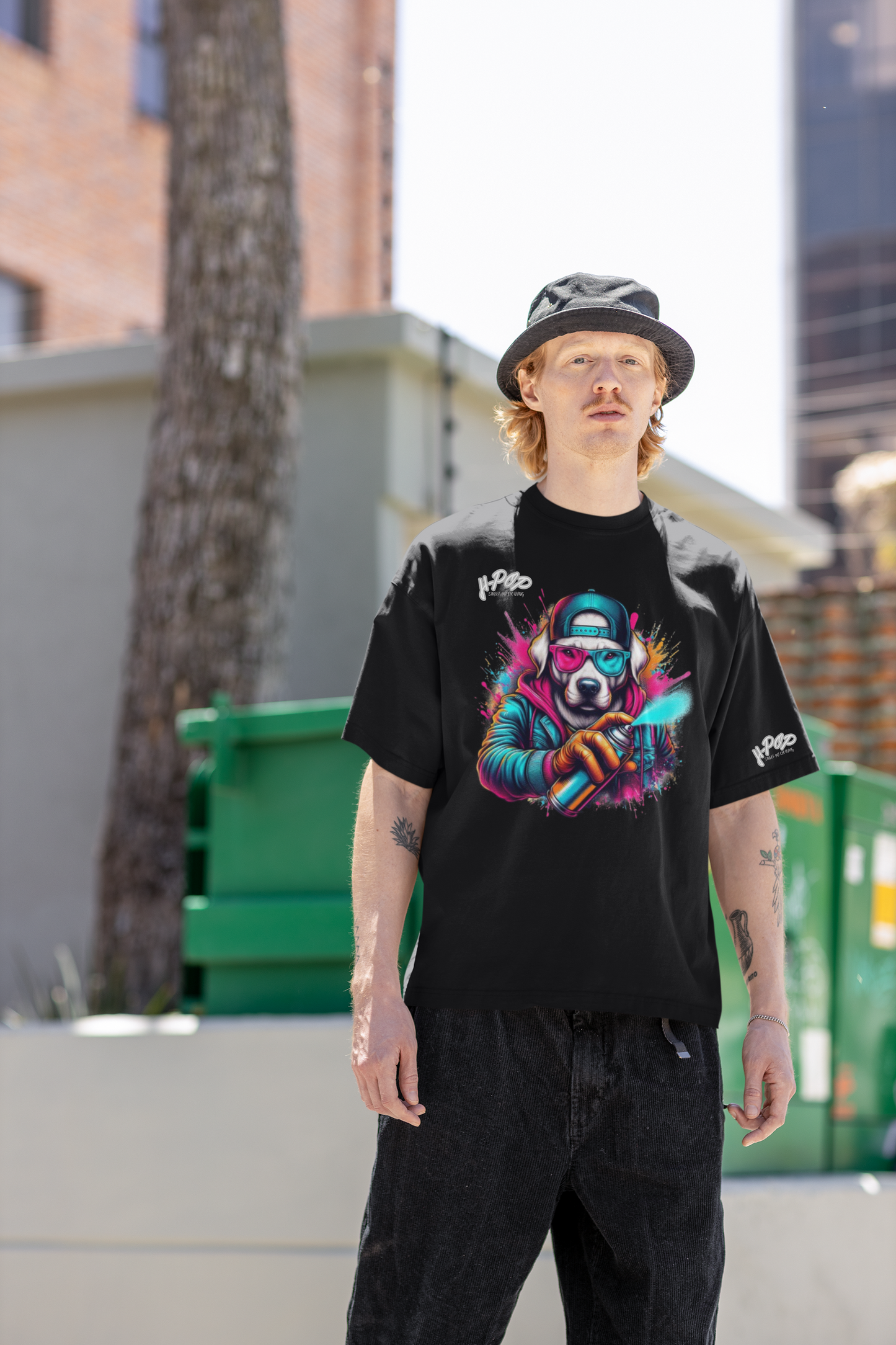 Un giovane uomo con un cappello da pescatore e capelli mossi indossa una maglietta nera oversize con una vivace stampa grafica colorata. La stampa raffigura un cane con un cappello, occhiali da sole rossi e blu, che tiene in mano una bomboletta spray con schizzi di colore. L'uomo è in piedi con le mani nelle tasche, con uno sguardo serio e posa in un ambiente urbano con un albero e un cassonetto verde sullo sfondo.