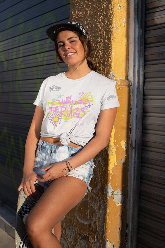 Una giovane donna sorridente con un cappellino da baseball scuro indossato all'indietro e lunghi capelli scuri, indossa una t-shirt bianca con una stampa grafica dai colori vivaci in stile neon o graffiti. La stampa sembra includere testo e un fiore stilizzato, con tonalità di rosa, giallo e verde lime. La maglietta è annodata in vita, mostrando l'ombelico, e la donna indossa pantaloncini di jeans corti. Si appoggia a un palo giallo consumato, con uno sfondo di una saracinesca metallica graffitata.
