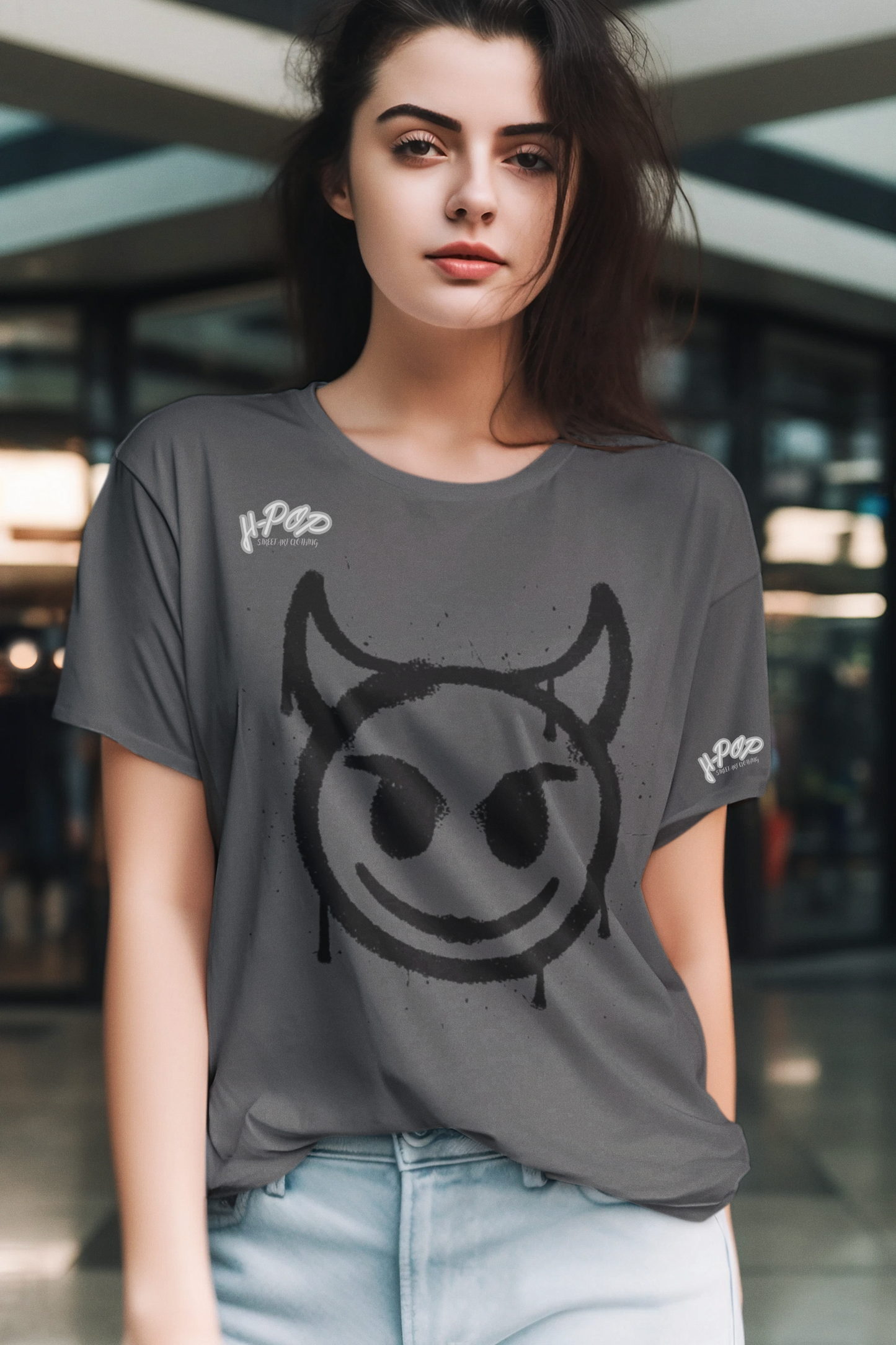 Una giovane donna indossa una t-shirt grigia casual con una stampa grafica centrale di una faccina sorridente stilizzata con corna, che ricorda un diavoletto o un'icona dallo stile underground, con un effetto spray. Indossa anche un paio di jeans chiari. Lo sfondo è sfocato e suggerisce un ambiente interno moderno.