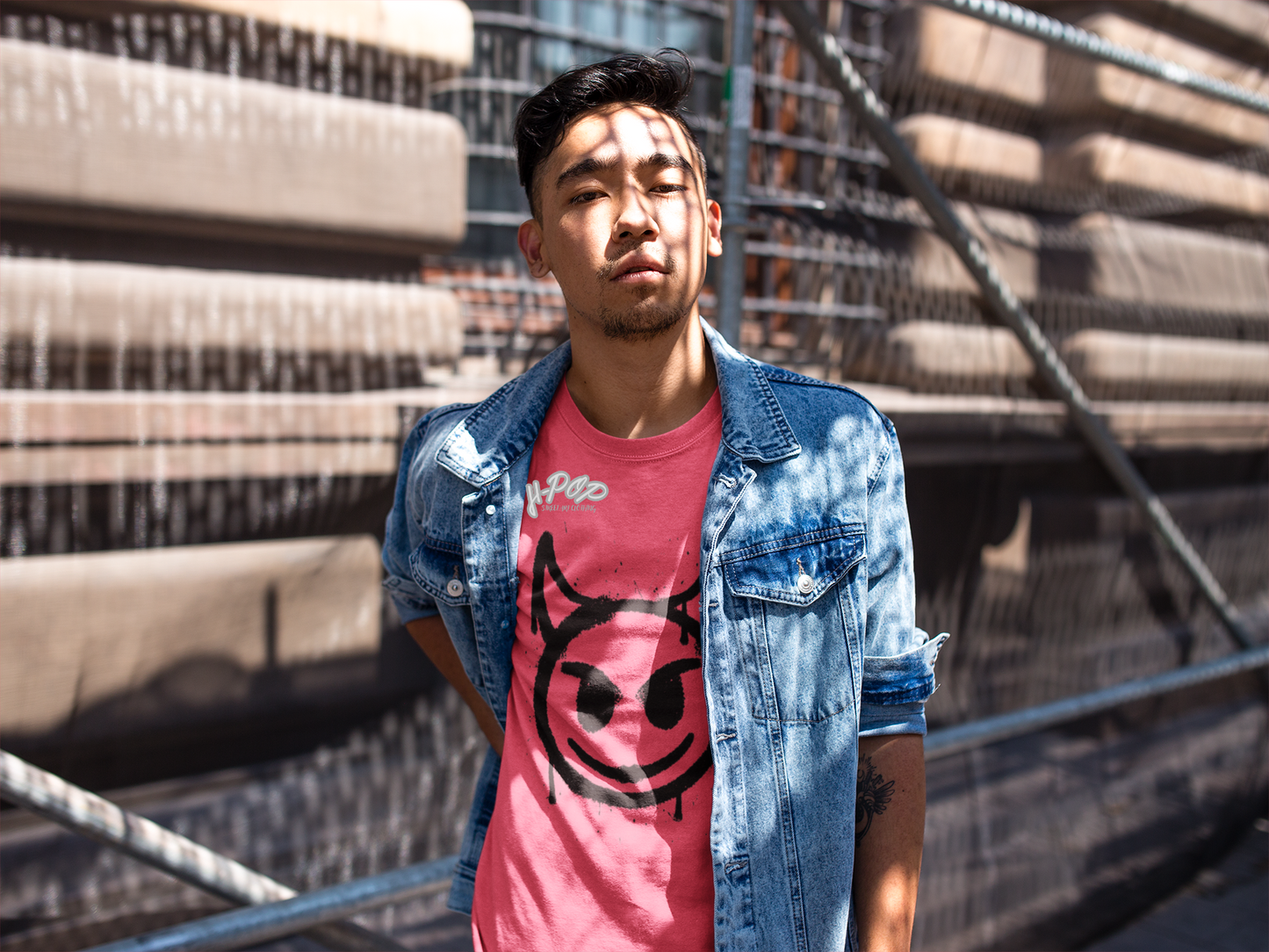 Un giovane uomo indossa una giacca di jeans chiara sopra una t-shirt rosa con una stampa grafica di una faccina sorridente stilizzata con corna, in stile diavoletto o graffito. Ha un'espressione seria e si trova in un ambiente urbano, con una struttura metallica e cemento sullo sfondo, e la luce del sole che crea ombre sul suo viso.