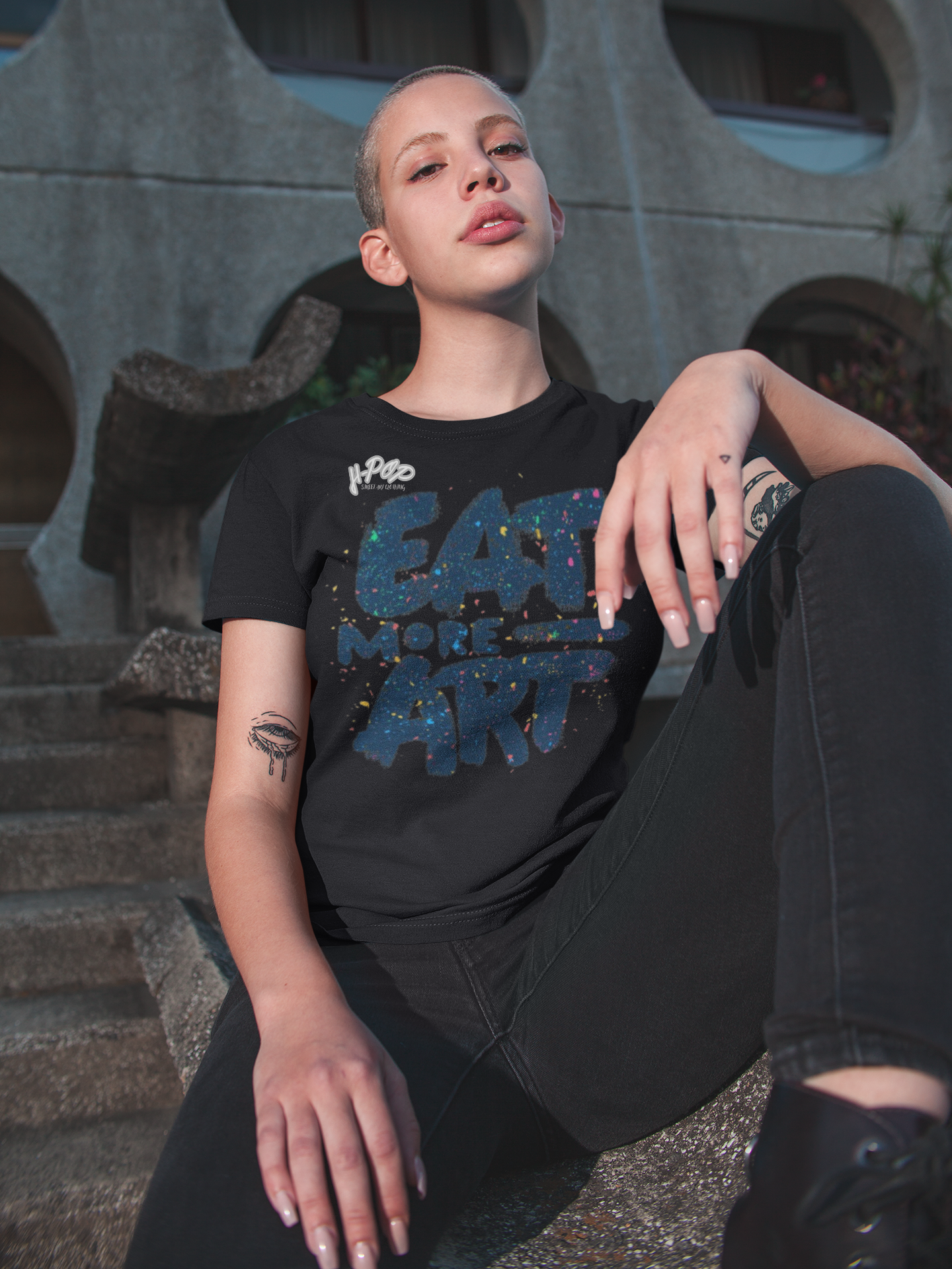 Donna con capelli rasati e tatuaggi, indossa una t-shirt nera con la scritta "EAT MORE ART" stilizzata con un effetto glitterato o "galaxy", abbinata a jeans scuri. Seduta su scalini di cemento, con uno sfondo architettonico. Perfetta per un look audace, artistico e street-chic. Parole chiave: t-shirt, maglietta, donna, moda, abbigliamento, arte, glitter, galaxy, casual, urban, street style, tatuaggi, tendenza, Italia.