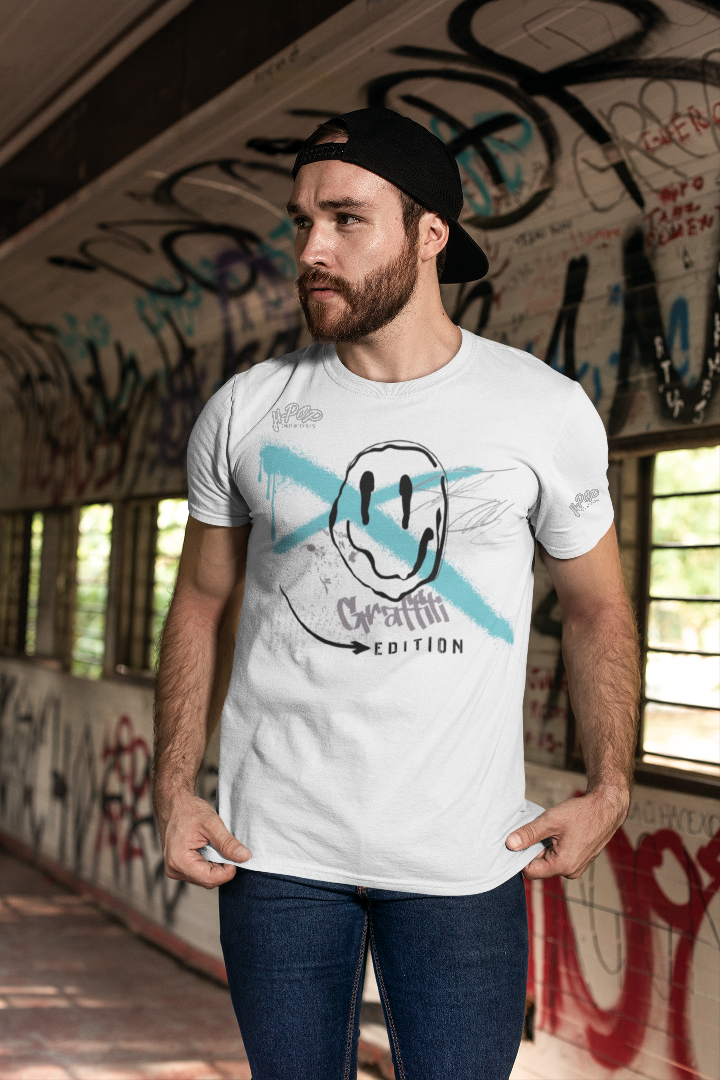 Giovane uomo con barba che indossa una t-shirt bianca con stampa stile graffiti, un cappellino nero all'indietro e jeans scuri, in un ambiente urbano decorato con graffiti. Parole chiave: t-shirt uomo, moda streetwear, abbigliamento urbano, t-shirt grafica, stile casual, moda italiana, maglietta graffiti, abbigliamento uomo, tendenze moda.