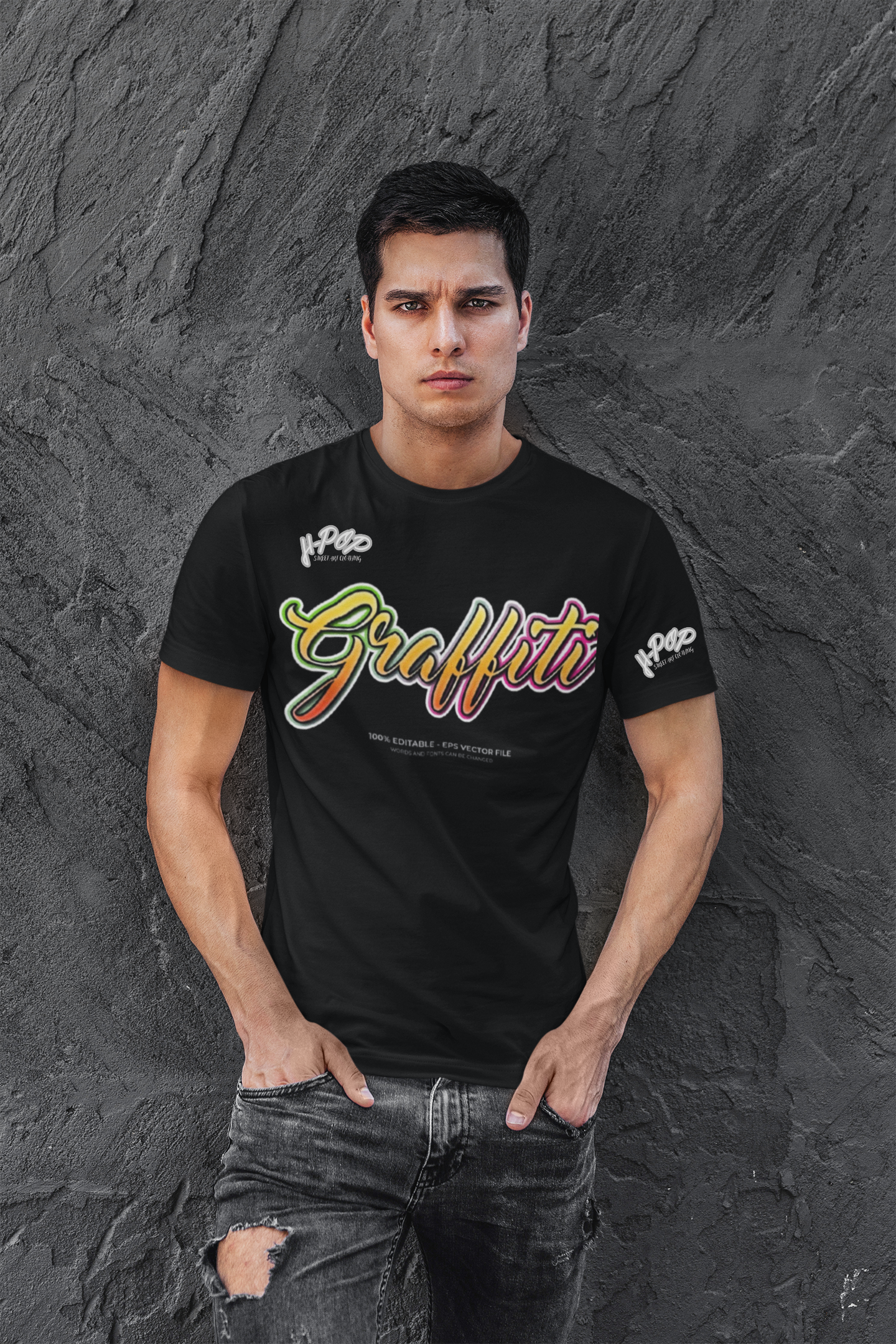 Un giovane uomo con un'espressione seria indossa una t-shirt nera con la scritta "Graffiti" in stile calligrafico dai colori vivaci e sfumati (giallo, arancione, rosso, rosa). Indossa jeans scuri strappati e tiene le mani nelle tasche, posando contro un muro scuro con una texture rocciosa o irregolare.
