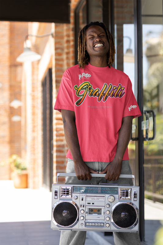 Un giovane uomo con dreadlocks e un'espressione divertita, quasi smorfiosa, indossa una t-shirt rosso corallo con la scritta "Graffiti" in stile calligrafico dai colori vivaci e sfumati (giallo, arancione, rosso). Regge un grande boombox vintage color argento con entrambe le mani. Si trova all'aperto, con uno sfondo che suggerisce l'ingresso di un edificio con mattoni e vetro.