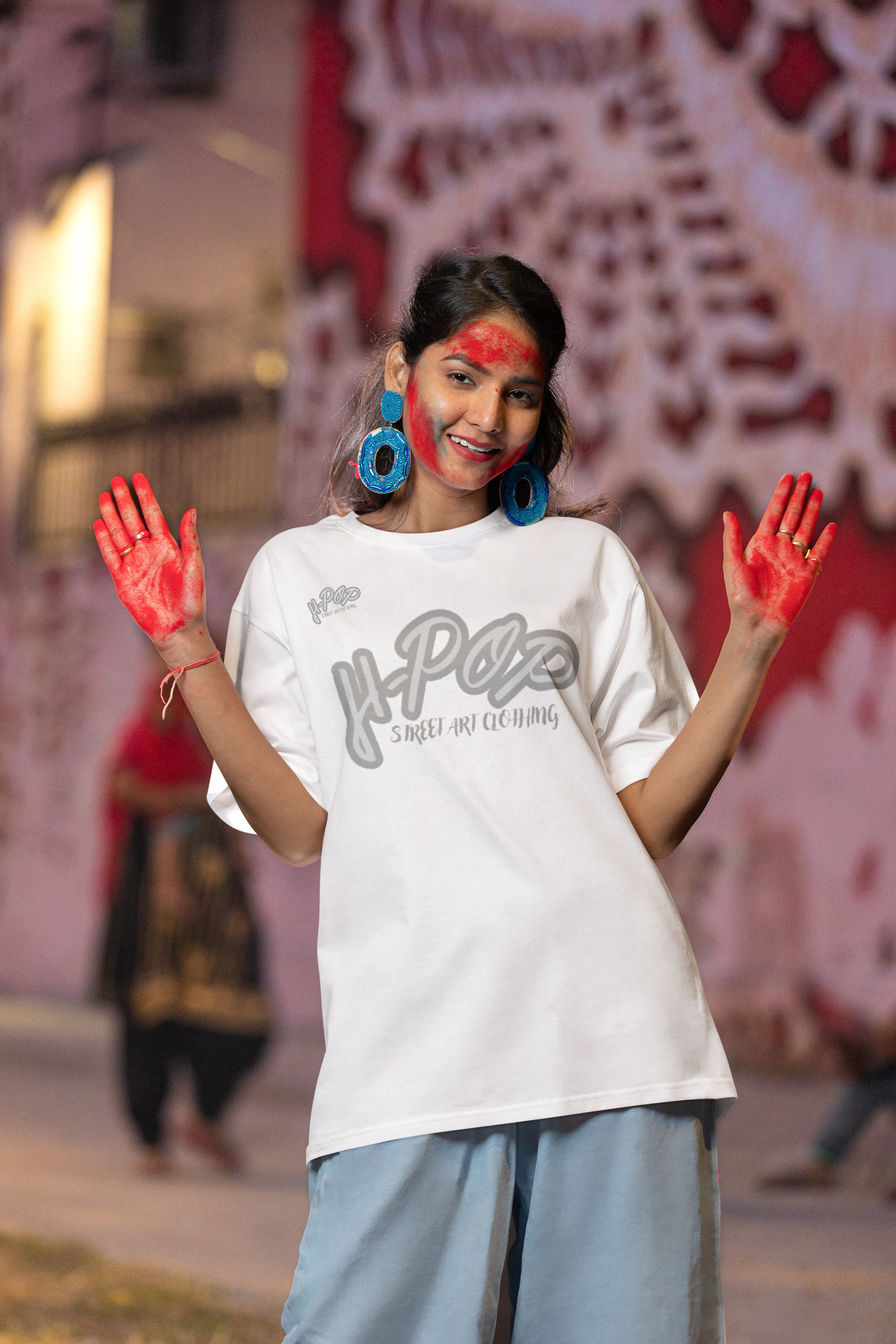 Una giovane donna sorridente, con del colore rosso sul viso e le mani dipinte di rosso, indossa grandi orecchini blu e una t-shirt bianca oversize con una scritta stilizzata grigia al centro che recita "H-Pop Street Art Clothing". Indossa pantaloni larghi di colore azzurro chiaro. Posa con le mani alzate e aperte, mostrando i palmi rossi, in un ambiente che suggerisce una strada o un vicolo, con luci soffuse e un muro decorato sullo sfondo. L'atmosfera è allegra e artistica.