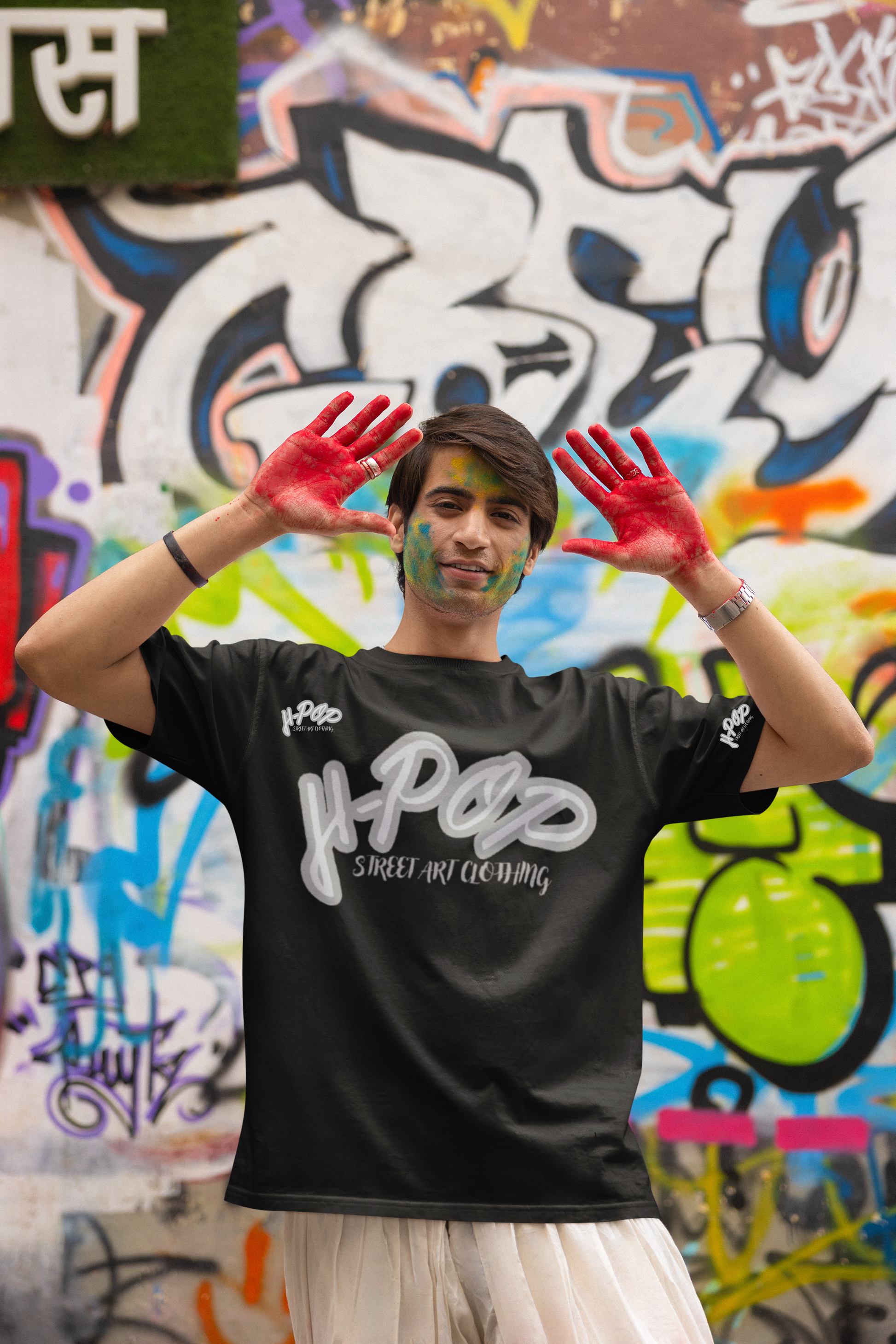 Un giovane uomo sorridente, con del colore sul viso e le mani dipinte di rosso, indossa una t-shirt nera con una scritta bianca stilizzata al centro che recita "H-Pop Street Art Clothing". Indossa pantaloni chiari e un orologio al polso. Posa con le mani alzate e aperte, mostrando i palmi rossi, davanti a un muro completamente coperto di graffiti colorati. L'espressione e il contesto suggeriscono un'atmosfera gioiosa e artistica.
