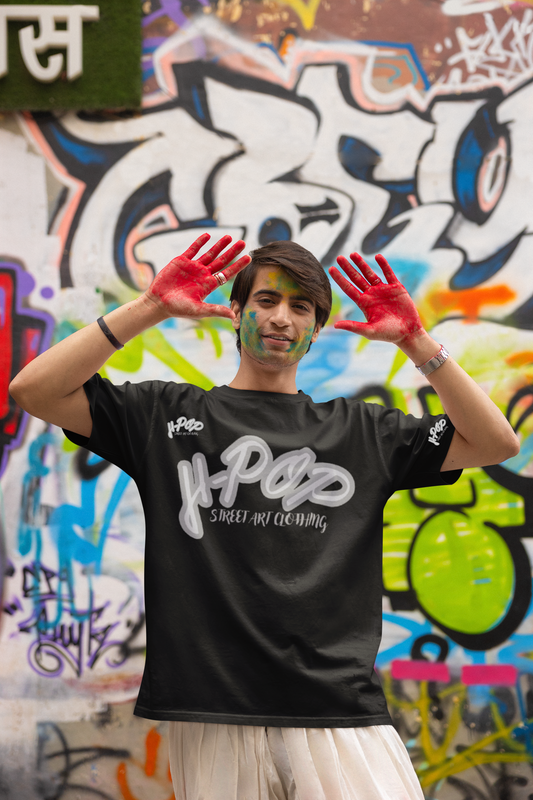 Un giovane uomo sorridente, con del colore sul viso e le mani dipinte di rosso, indossa una t-shirt nera con una scritta bianca stilizzata al centro che recita "H-Pop Street Art Clothing". Indossa pantaloni chiari e un orologio al polso. Posa con le mani alzate e aperte, mostrando i palmi rossi, davanti a un muro completamente coperto di graffiti colorati. L'espressione e il contesto suggeriscono un'atmosfera gioiosa e artistica.