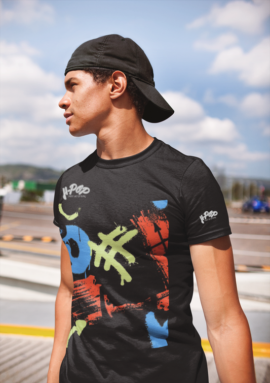 Un giovane uomo con un cappellino da baseball nero indossato all'indietro indossa una t-shirt nera con una stampa grafica astratta e colorata sul davanti. La stampa presenta forme e tratti che ricordano graffiti o pennellate di colori vivaci come blu, arancione e verde lime. Si trova all'aperto, in un ambiente che suggerisce una strada o un'area urbana con un cielo nuvoloso sullo sfondo.