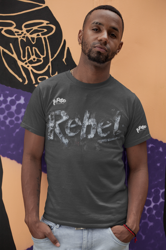 Un giovane uomo con barba curata e piercing all'orecchio indossa una t-shirt grigio antracite con la scritta "Rebel" stampata in stile graffito o consumato, con un effetto ruvido e quasi tridimensionale. Indossa jeans chiari e tiene le mani nelle tasche. Posa con un leggero sorriso, inclinato, davanti a un muro decorato con murales stilizzati in tonalità scure e viola su sfondo arancione.