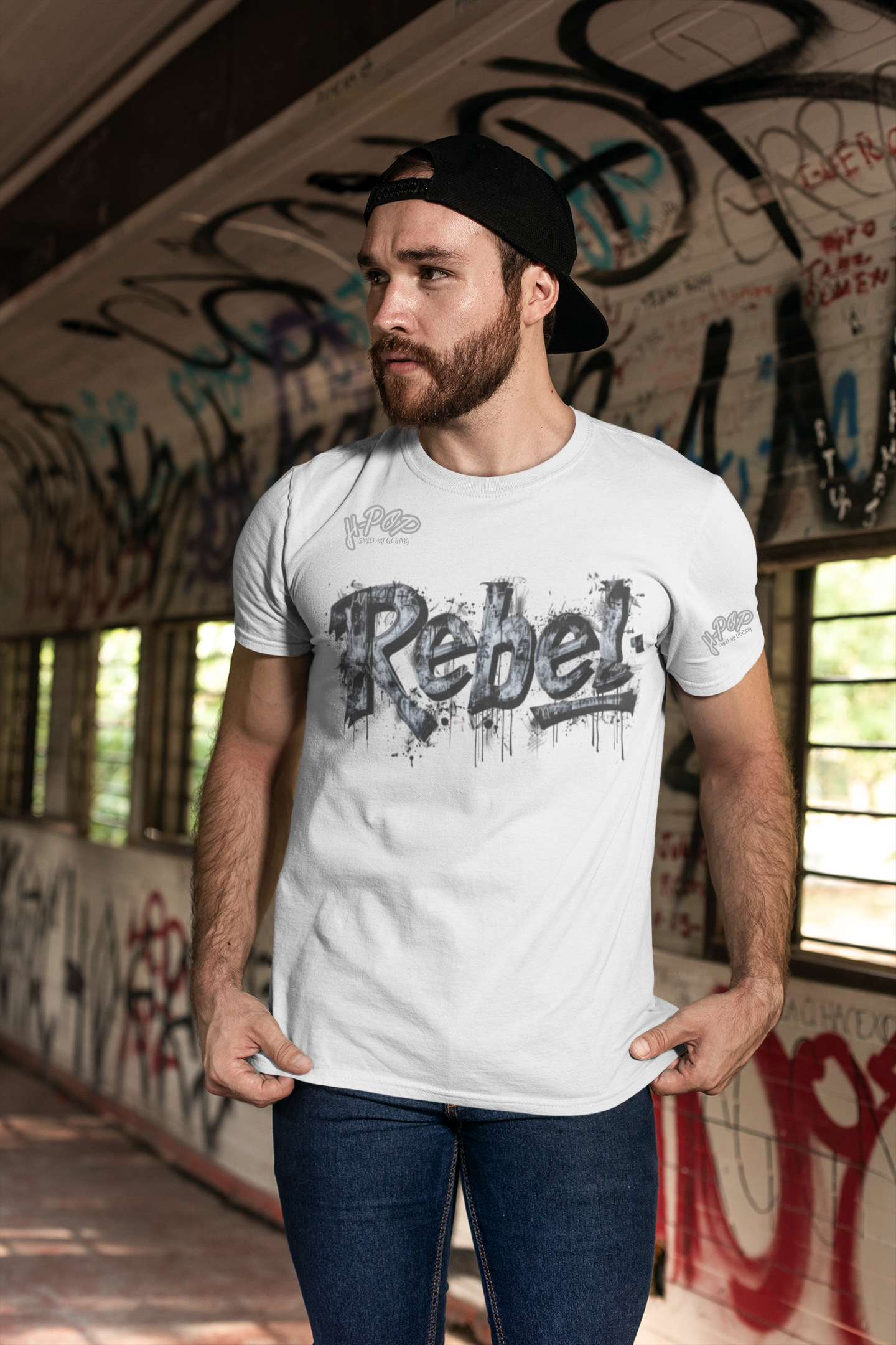 Un giovane uomo con barba e un cappellino da baseball nero indossato all'indietro indossa una t-shirt bianca con la scritta "Rebel" in stile graffito, con un effetto sporco e gocciolante. Indossa jeans scuri e posa con le mani che tirano leggermente la maglietta, guardando di lato. Si trova in un ambiente che ricorda un sottopassaggio o un tunnel, con muri ricoperti di graffiti.