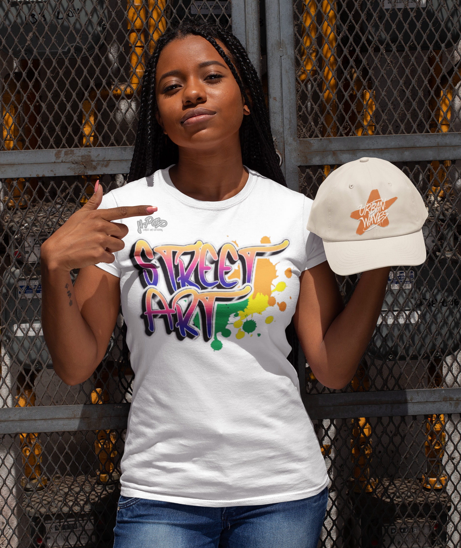 Una giovane donna con lunghe treccine indossa una t-shirt bianca con una stampa grafica vibrante che recita "STREET ART" in stile graffiti, con colori sfumati e schizzi. Con una mano indica la maglietta, mentre nell'altra tiene un cappellino beige con un logo arancione stilizzato. Posa davanti a una recinzione metallica scura, con uno sfondo urbano.