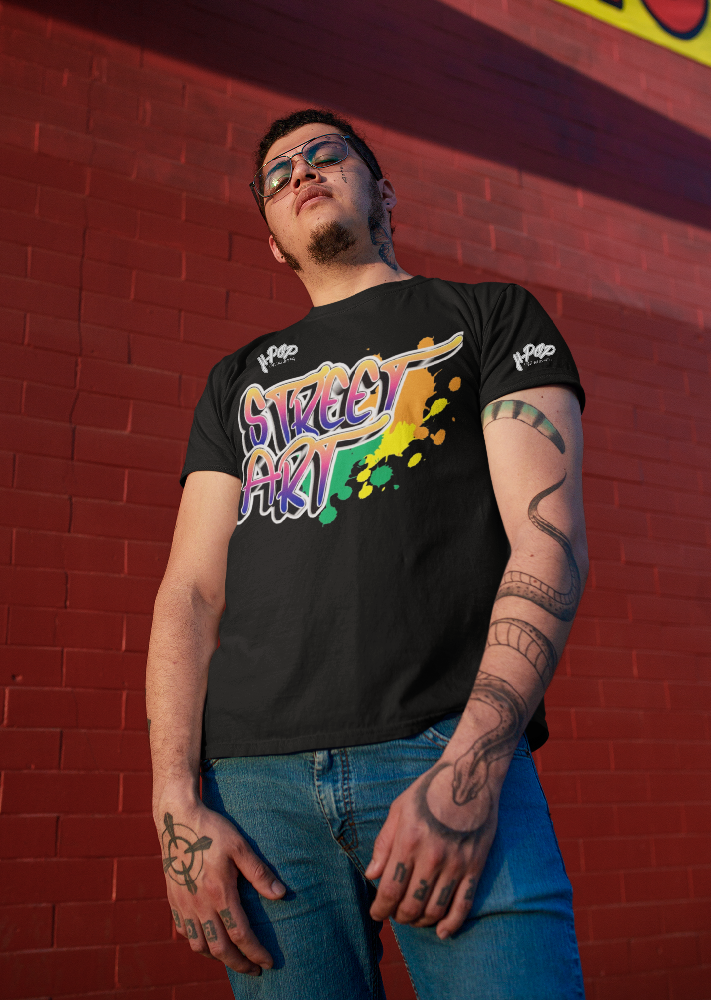 Un giovane uomo con occhiali e tatuaggi sulle braccia e sulle mani indossa una t-shirt nera con una vivace stampa grafica "STREET ART" in stile graffiti, con colori sfumati e schizzi. Indossa jeans e posa guardando verso l'alto, con uno sfondo di un muro di mattoni rossi e luci gialle.