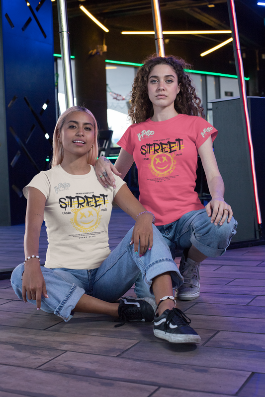 Due ragazze giovani indossano t-shirt casual con grafica "STREET" e faccina sorridente, abbinate a jeans ampi. Una ragazza ha una maglietta color crema e l'altra una maglietta rosa corallo. Sono sedute su un pavimento in legno con luci al neon sfocate sullo sfondo, in un ambiente che suggerisce un evento o un locale notturno.