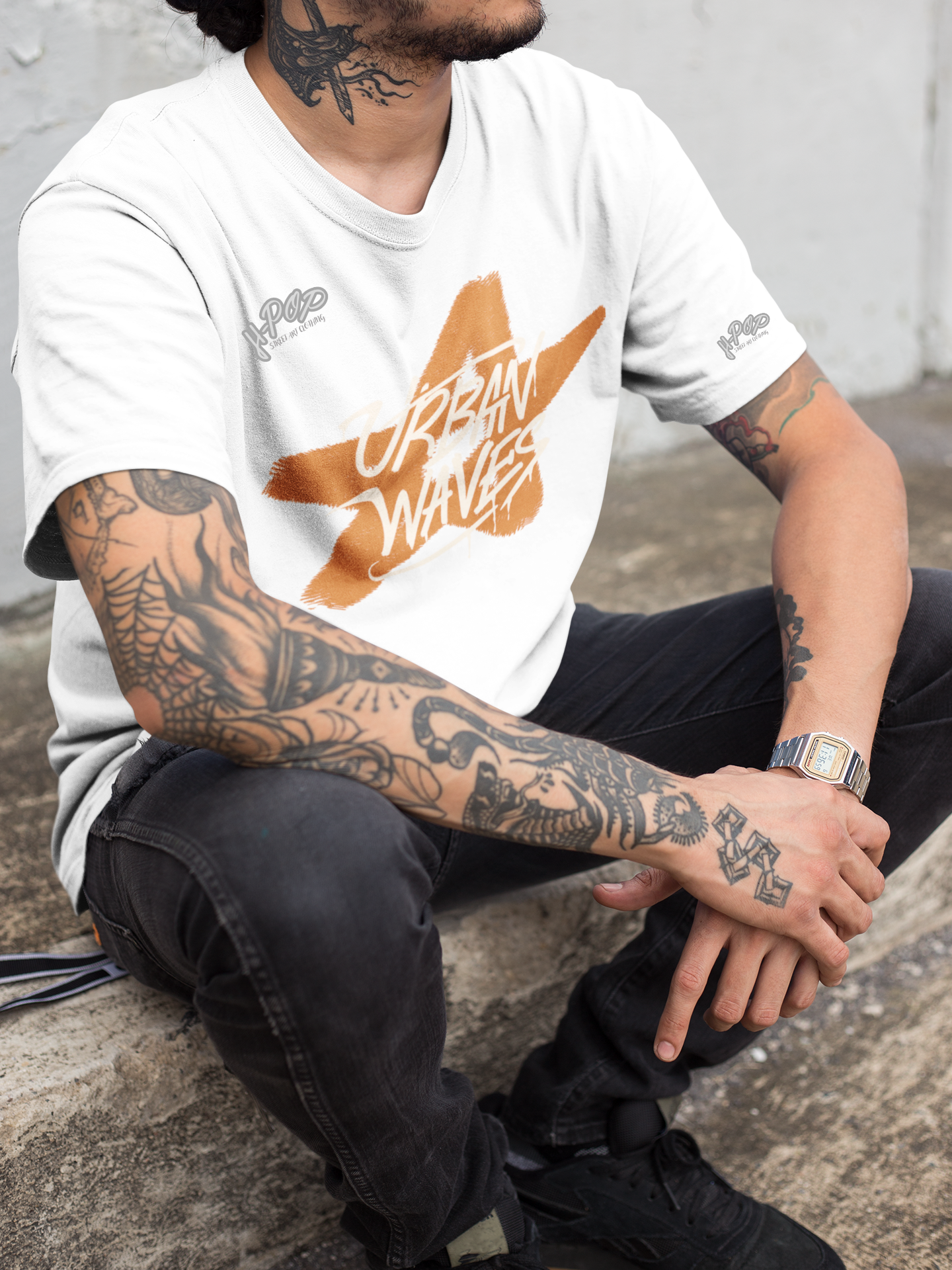 Primo piano di un uomo con tatuaggi su braccia e collo, che indossa una t-shirt bianca con una stampa arancione di una stella e la scritta "URBAN WAVES". Un orologio al polso sinistro e jeans scuri completano il look casual. Parole chiave: t-shirt, maglietta, uomo, moda, abbigliamento, streetwear, casual, urban, tatuaggi, stella, design, stile, tendenza, Italia.