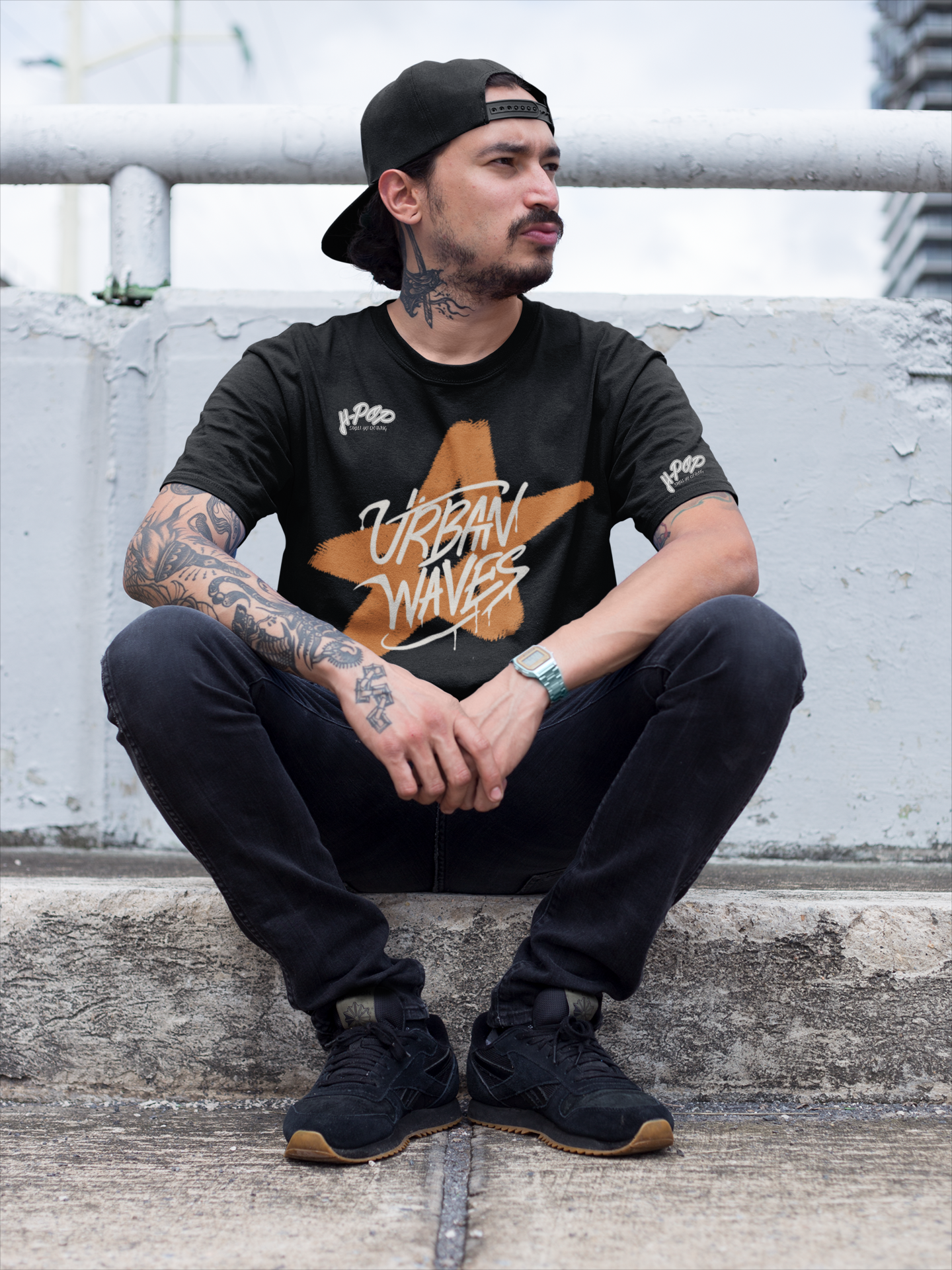 Uomo con tatuaggi e cappellino all'indietro, indossa una t-shirt nera con una stella arancione stilizzata e la scritta "URBAN WAVES", jeans scuri e sneakers nere. Seduto su gradini in cemento. Perfetto per un look streetwear e casual. Parole chiave: t-shirt, maglietta, uomo, moda, abbigliamento, streetwear, casual, urban, tatuaggi, stella, design, stile, tendenza, Italia.