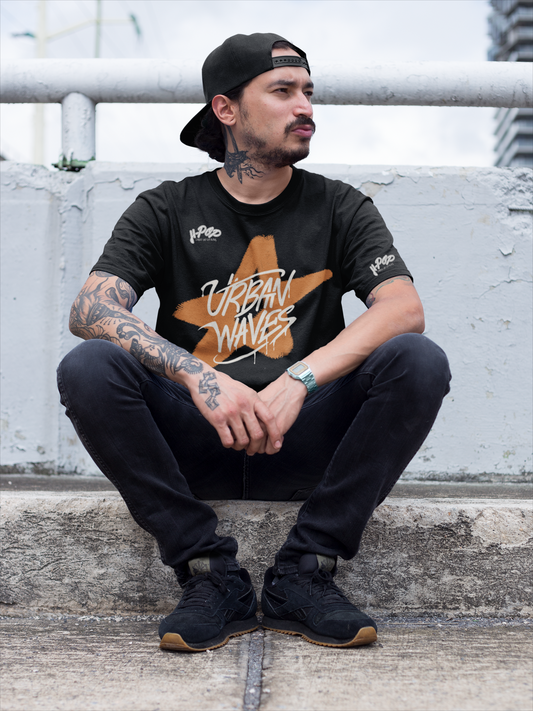 Uomo con tatuaggi e cappellino all'indietro, indossa una t-shirt nera con una stella arancione stilizzata e la scritta "URBAN WAVES", jeans scuri e sneakers nere. Seduto su gradini in cemento. Perfetto per un look streetwear e casual. Parole chiave: t-shirt, maglietta, uomo, moda, abbigliamento, streetwear, casual, urban, tatuaggi, stella, design, stile, tendenza, Italia.