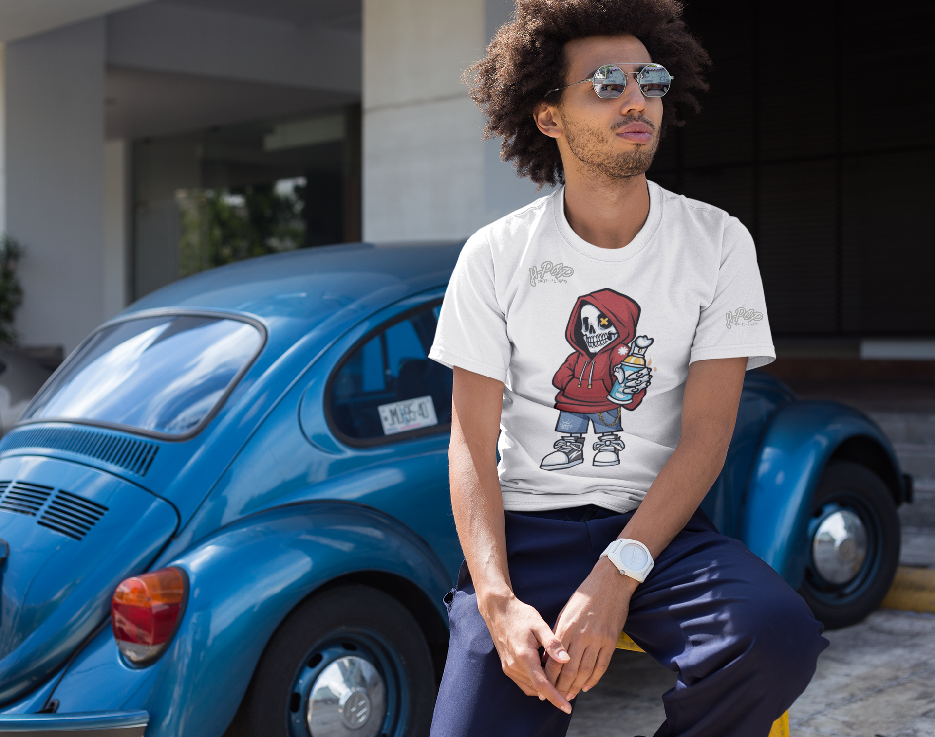 Un giovane uomo con capelli ricci e occhiali da sole tondi indossa una maglietta bianca con una stampa grafica di un personaggio scheletro cartoonesco che indossa una felpa con cappuccio rossa e jeans, con una bomboletta spray in mano e una catena. Lo scheletro ha un occhio a "X". La maglietta ha anche il logo "H-Pop" in alto a sinistra e sulla manica. L'uomo è seduto su un parapetto giallo accanto a un Maggiolino Volkswagen blu d'epoca, guardando di lato.