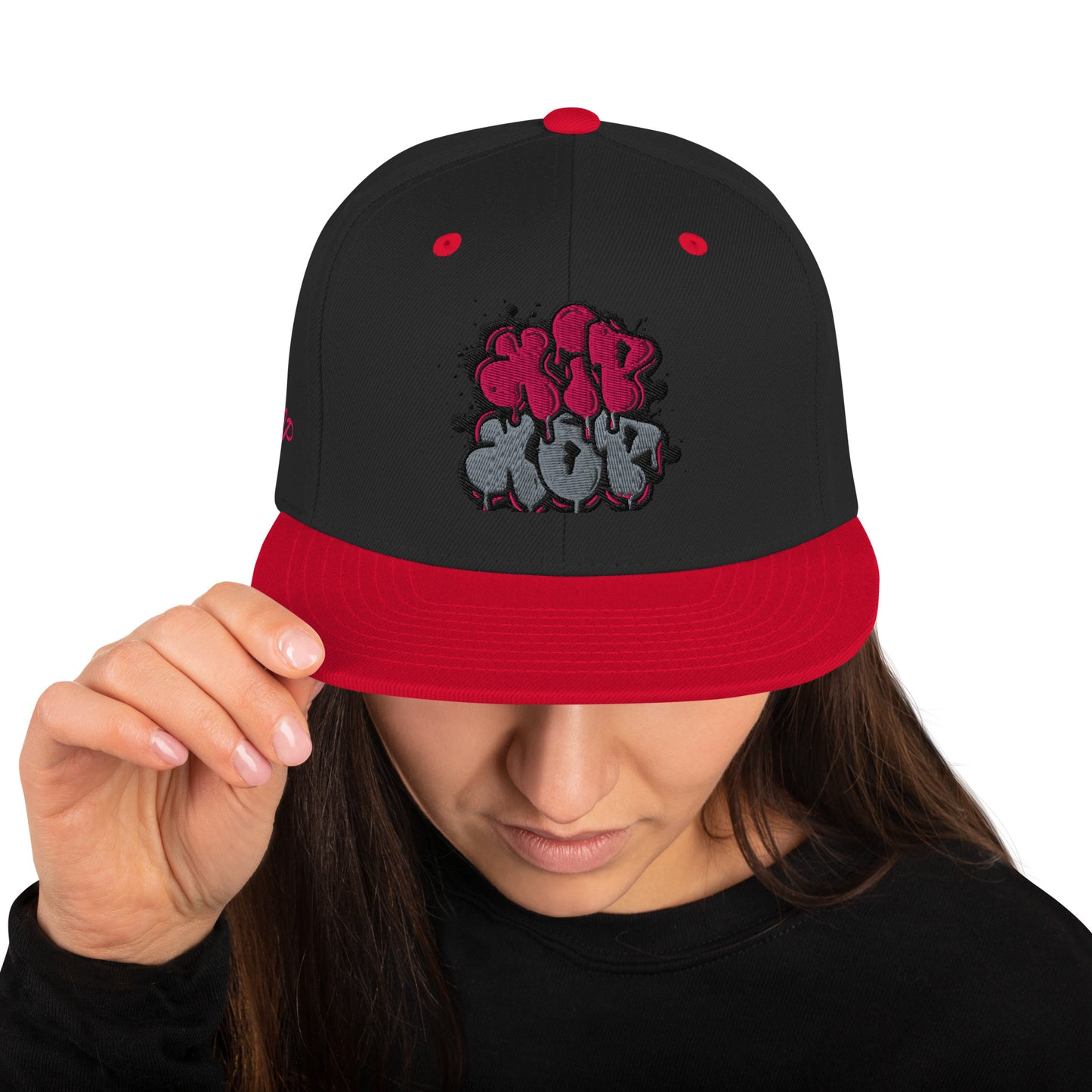 Una donna con i capelli scuri che indossa un cappellino snapback nero con visiera rossa. Il cappellino presenta una stampa grafica in stile graffiti con la scritta 'Hip Hop' in tonalità rosa e grigie. Ideale per un look streetwear e casual. Parole chiave: cappellino, cappellino snapback, cappellino Hip Hop, streetwear, moda casual, accessori moda, cappellino nero e rosso, stile graffiti, abbigliamento H-Pop, tendenze moda Italia.