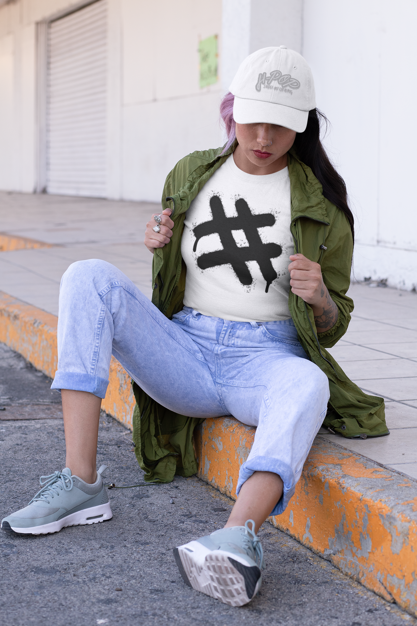 Una giovane donna indossa un cappellino beige, una t-shirt bianca con una grande stampa nera in stile graffito raffigurante un simbolo di hashtag (#) con effetto gocciolante. Sopra la t-shirt indossa una giacca verde militare aperta. Completa il look con jeans chiari arrotolati alle caviglie e scarpe da ginnastica. È seduta sul bordo di un marciapiede, con le mani che tengono il bavero della giacca.