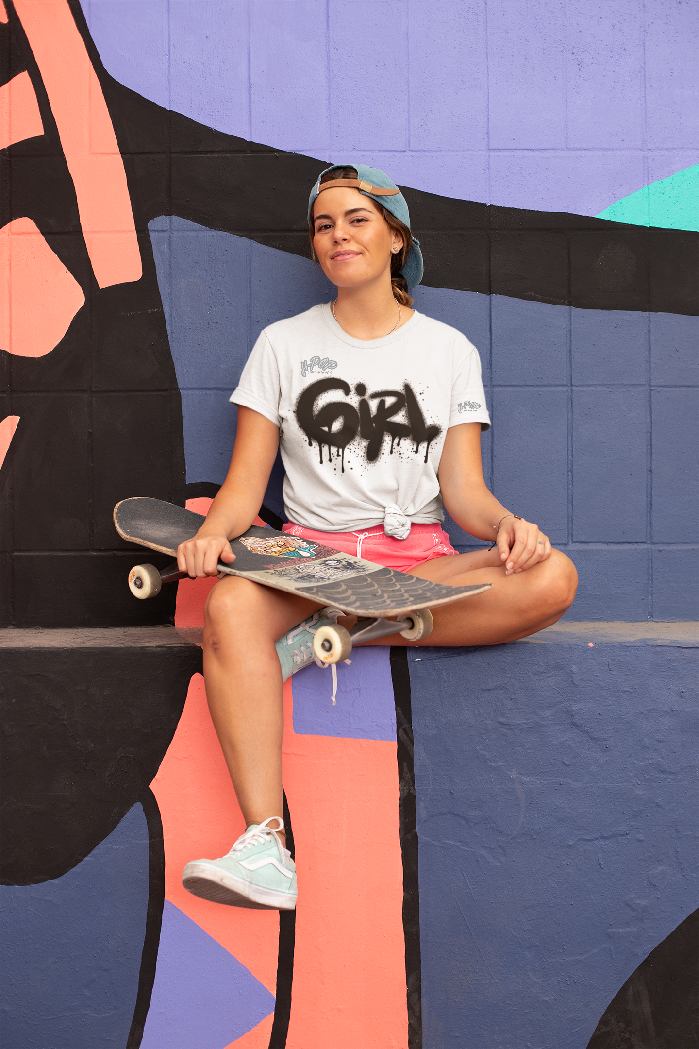 Una giovane donna con un berretto girato all'indietro e una t-shirt bianca con la scritta "GIRL" in stile graffito nero, con effetto gocciolante, siede con le gambe incrociate su un bordo di cemento colorato. Tiene uno skateboard appoggiato alle gambe e guarda sorridendo verso la fotocamera. Lo sfondo è un muro con murales astratti nei toni del viola, arancione e nero.