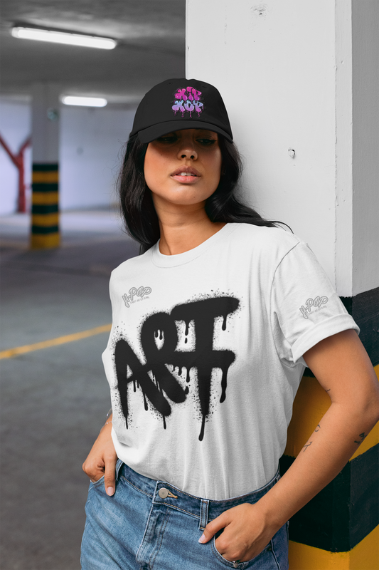 Ragazza con cappellino nero e t-shirt bianca con stampa "ART" in stile graffiti dripping, in un parcheggio sotterraneo. Ideale per un look streetwear e artistico. Parole chiave: t-shirt, maglietta, streetwear, moda, abbigliamento, donna, cappellino, graffiti, arte, casual, urban, stile, tendenza, Italia.