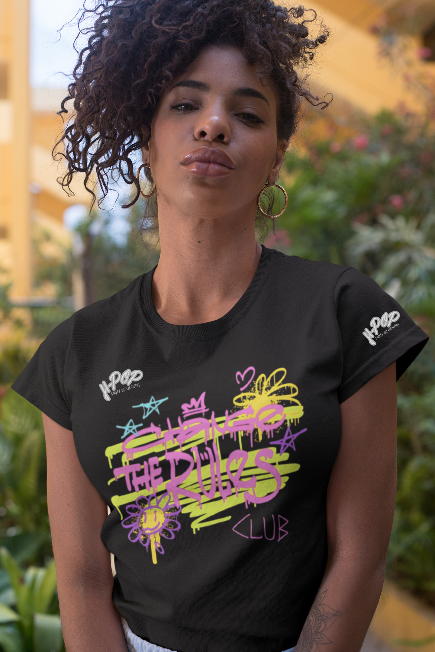 Una giovane donna con capelli ricci scuri e orecchini a cerchio indossa una t-shirt nera con una stampa grafica vibrante e in stile graffiti. La stampa presenta scritte stilizzate e simboli come stelle, corone e fiori, in colori neon come il giallo lime, il rosa e il viola. Posa guardando direttamente la fotocamera, con uno sfondo sfocato che suggerisce un ambiente esterno con vegetazione e architettura.