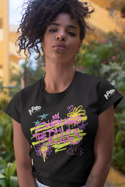 Una giovane donna con capelli ricci scuri e orecchini a cerchio indossa una t-shirt nera con una stampa grafica vibrante e in stile graffiti. La stampa presenta scritte stilizzate e simboli come stelle, corone e fiori, in colori neon come il giallo lime, il rosa e il viola. Posa guardando direttamente la fotocamera, con uno sfondo sfocato che suggerisce un ambiente esterno con vegetazione e architettura.