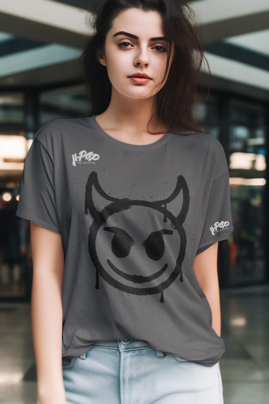 Una giovane donna indossa una t-shirt grigia casual con una stampa grafica centrale di una faccina sorridente stilizzata con corna, che ricorda un diavoletto o un'icona dallo stile underground, con un effetto spray. Indossa anche un paio di jeans chiari. Lo sfondo è sfocato e suggerisce un ambiente interno moderno. 