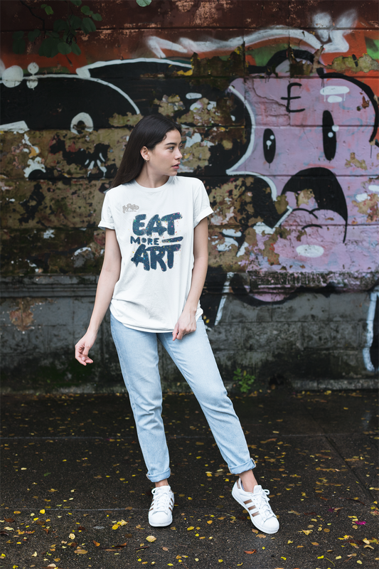 Ragazza con t-shirt bianca con la scritta "EAT MORE ART" in stile graffiti blu scuro, jeans chiari e sneakers bianche, in piedi davanti a un muro coperto di murales. Perfetta per un look casual, artistico e urbano. Parole chiave: t-shirt, maglietta, donna, moda, abbigliamento, arte, graffiti, casual, urban, street style, tendenza, Italia.