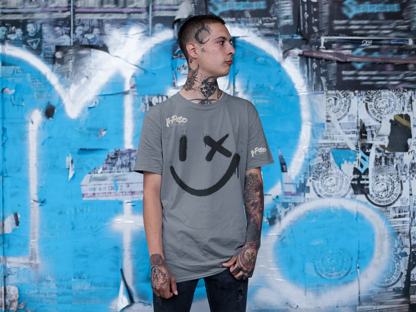 Un giovane uomo con tatuaggi sul collo e sulle braccia indossa una maglietta grigia con una grande stampa nera di uno smiley stilizzato: un occhio è un tratto verticale e l'altro una "X", con un effetto vernice spray. La maglietta ha anche il logo "H-Pop" in alto a sinistra e sulla manica. L'uomo è in piedi con le mani nelle tasche e guarda di lato, posando davanti a un muro coperto di graffiti e poster.