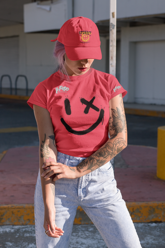 Una giovane donna con capelli rosa e braccia tatuate indossa una maglietta rosso corallo con una grande stampa nera di uno smiley stilizzato: un occhio è un tratto verticale e l'altro una "X". Indossa anche un cappellino da baseball rosso con la scritta "STREET". La donna è in piedi all'aperto, con lo sguardo abbassato, in un contesto urbano.