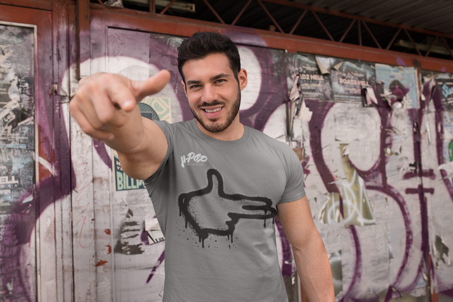 Un uomo sorridente con una maglietta grigia stampata con un'illustrazione "pistola con le dita" stilizzata. Si trova davanti a un muro di legno con graffiti e poster. L'uomo punta un pollice verso l'alto con un sorriso radioso, trasmettendo un'atmosfera positiva ed energica.