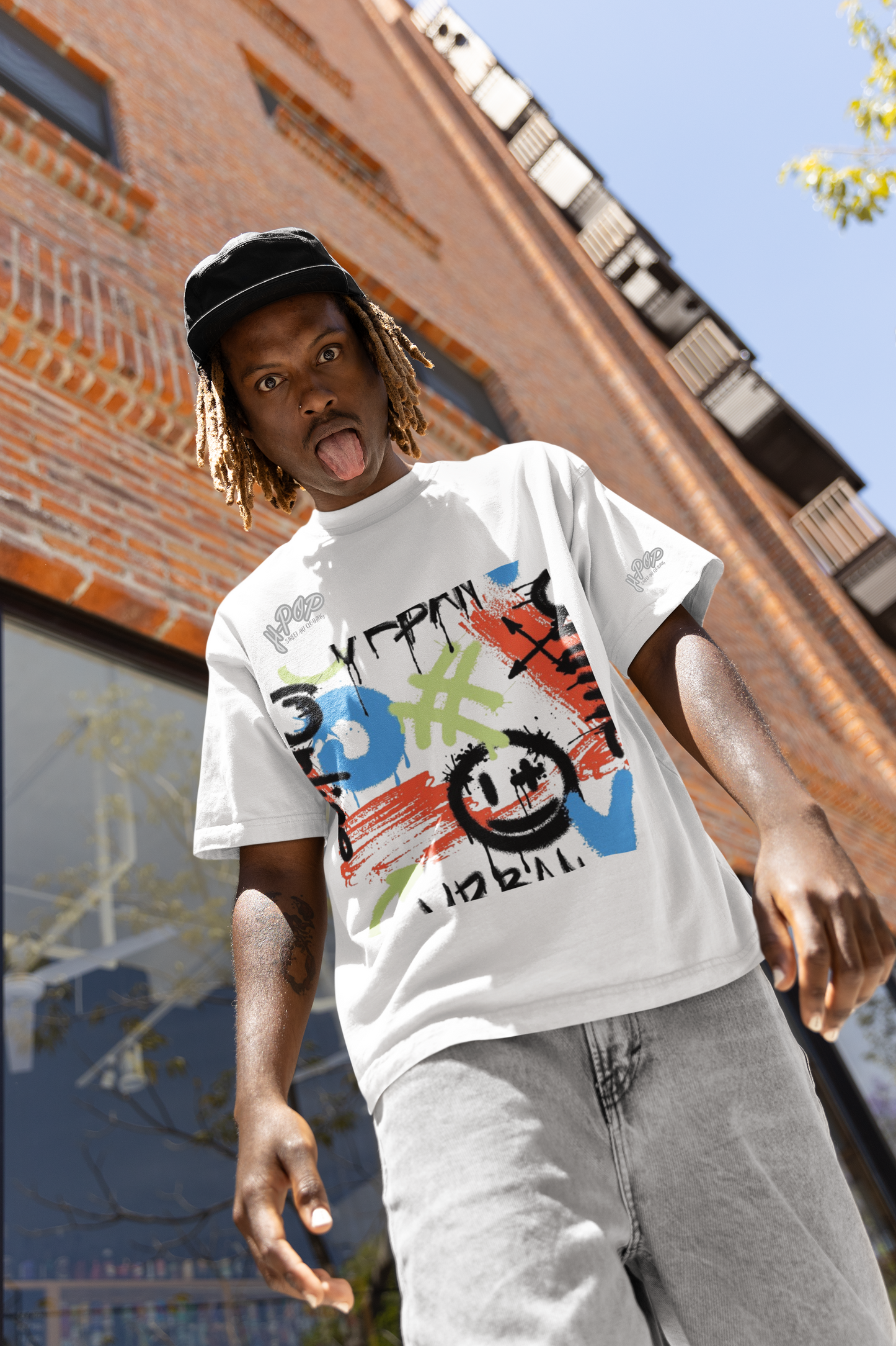 Un giovane uomo con treccine chiare e un cappellino nero indossa una t-shirt bianca con una vivace stampa grafica in stile graffiti. La stampa include forme e simboli astratti in colori come blu, rosso, verde lime e nero, con una faccina sorridente stilizzata. Indossa jeans chiari e tira fuori la lingua verso la fotocamera. Si trova all'aperto, con un edificio in mattoni e un cielo sereno sullo sfondo.