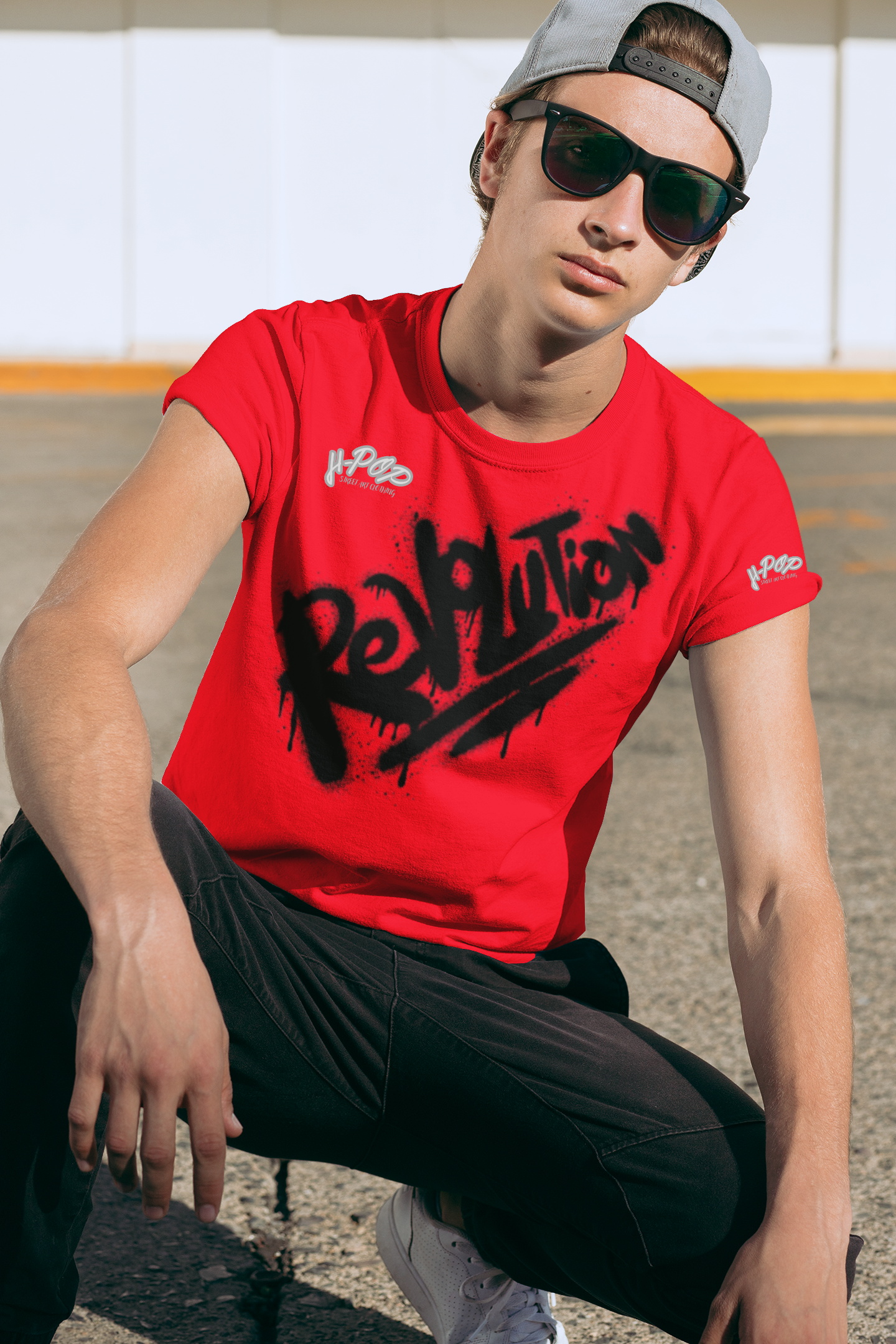 Giovane uomo che indossa una maglietta rossa con stampa 'Revolution' in stile graffiti, pantaloni neri, occhiali da sole e cappellino all'indietro. Look streetwear energico e abbigliamento urbano maschile. Scopri la nostra selezione di t-shirt stampate e moda giovanile.