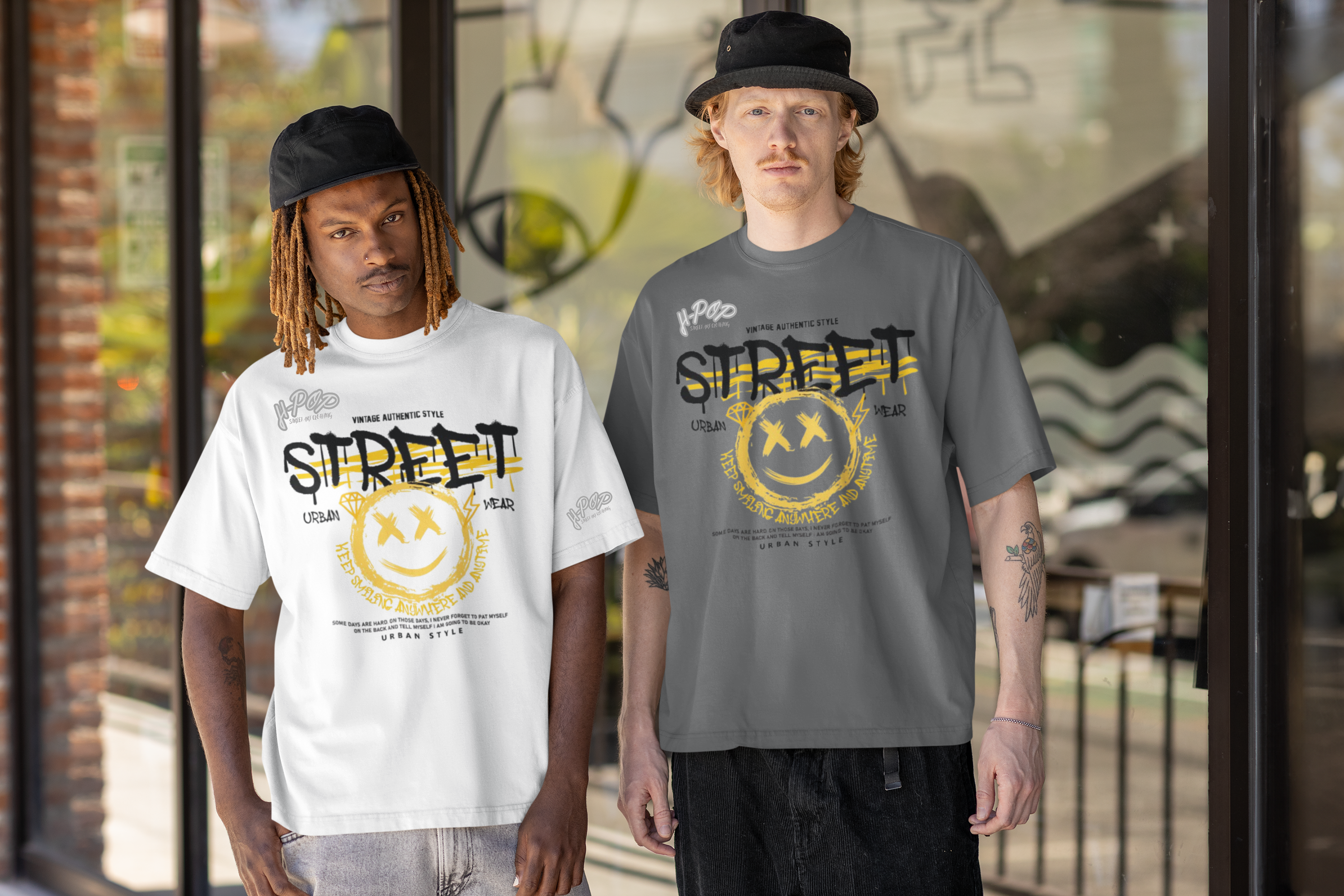 Due uomini indossano t-shirt oversize con un design audace che include la parola "STREET" e una faccina sorridente graffiata. Uno indossa una maglietta bianca e l'altro una grigia, entrambi con cappelli casual e posano in un ambiente urbano, forse di fronte a un negozio o un edificio moderno. 