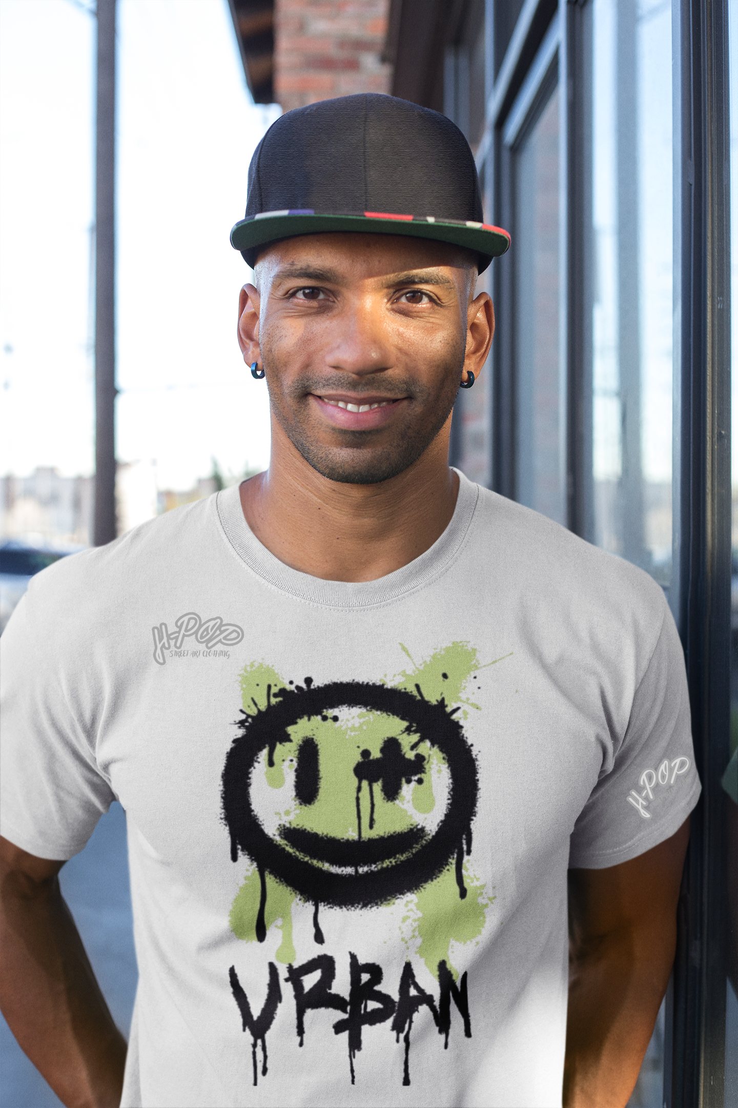 Uomo sorridente che indossa una maglietta grigio chiaro con stampa 'Urban' e faccina sorridente effetto graffiti verde e nero. Completa il look con un cappellino scuro e orecchini. Ideale per abbigliamento streetwear maschile e moda urbana casual.