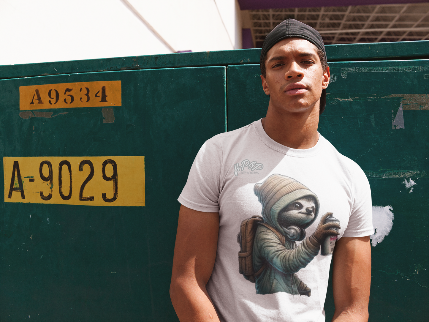 Un giovane uomo muscoloso con un cappellino da baseball nero indossato all'indietro indossa una t-shirt bianca con una stampa che raffigura un bradipo vestito con un cappuccio e uno zaino, mentre tiene in mano una bomboletta spray, come se stesse facendo graffiti. Il bradipo ha un'espressione divertita. L'uomo posa con un'espressione seria, appoggiandosi a una parete verde scuro con scritte e numeri gialli.
