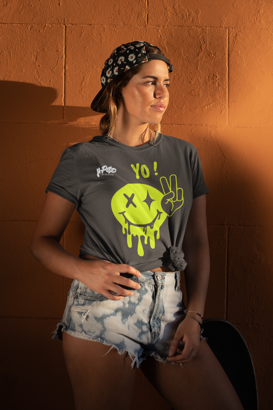 Una giovane donna con un cappellino da baseball fantasia indossato all'indietro e una maglietta grigio scuro annodata in vita. La maglietta presenta una grande grafica verde lime di uno smiley stilizzato e gocciolante, con un occhio a "X" e l'altro una "X" con due stelle, e accanto la scritta "YO!". Indossa anche pantaloncini di jeans chiari strappati. La donna è in piedi appoggiata a un muro di mattoni marroni, con uno skateboard parzialmente visibile accanto a lei.