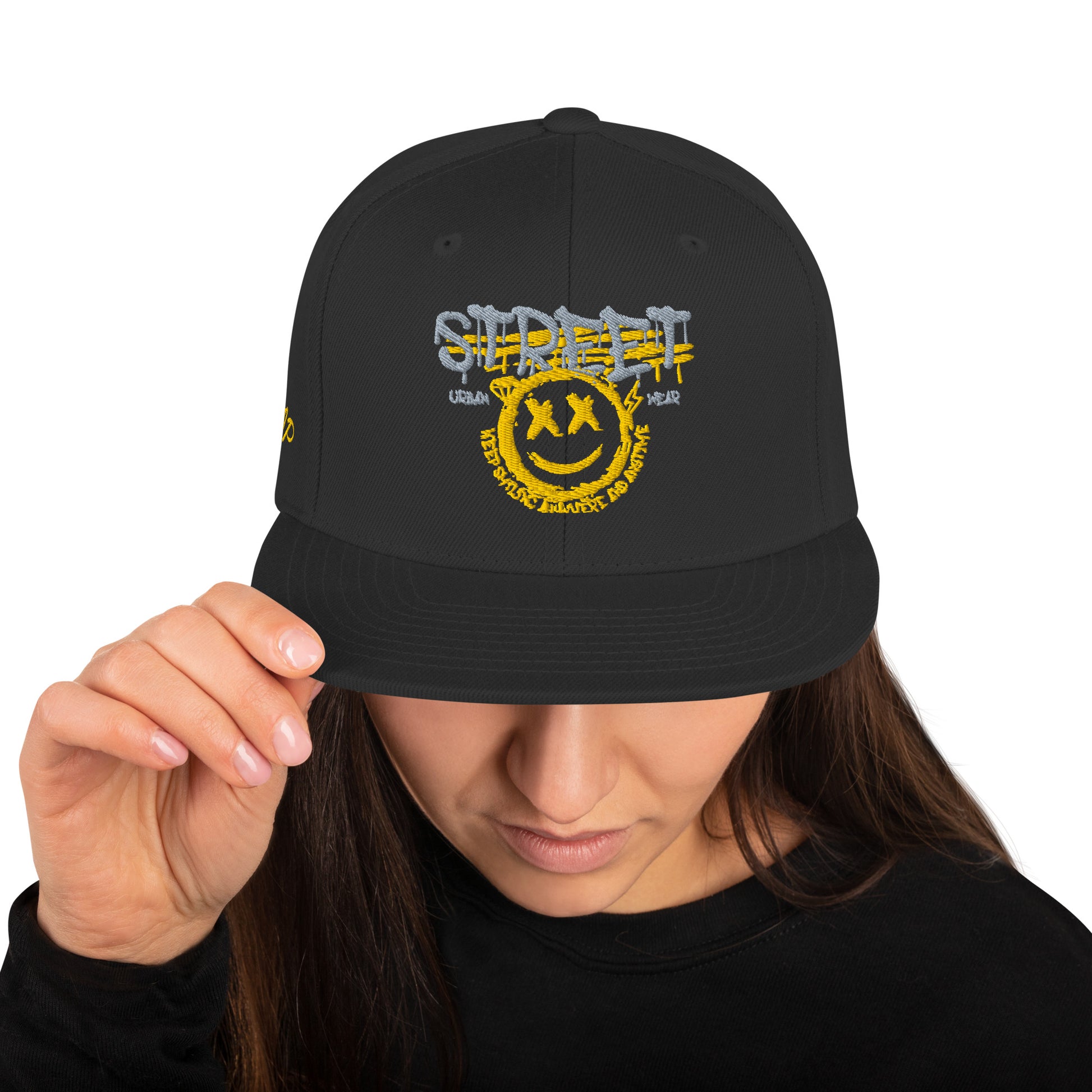 Donna che indossa un cappellino snapback nero con ricamo in stile graffiti: "STREETWEAR" in grigio e giallo, un volto sorridente giallo con occhi a "X" e la scritta circolare "KEEP SMILING BELIEVE AND SHINE". Ideale per abbigliamento urbano e accessori di tendenza. Parole chiave: cappellino, snapback, streetwear, graffiti, ricamo, moda donna, accessori, urban style, smile, nero, giallo, tendenza, Italia.