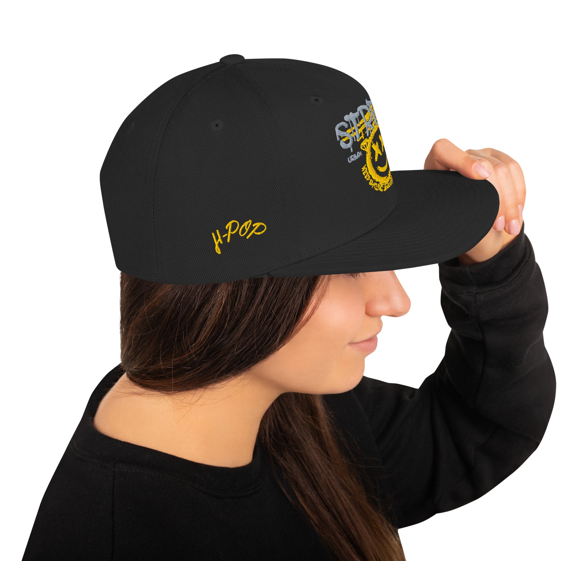 Donna di profilo che indossa un cappellino snapback nero con ricamo in stile graffiti: "STREETWEAR" in grigio e giallo, un volto sorridente giallo con occhi a "X" e la scritta circolare "KEEP SMILING BELIEVE AND SHINE" sul davanti, e il logo "H-Pop" ricamato in giallo sul lato. Ideale per abbigliamento urbano e accessori di tendenza. Parole chiave: cappellino, snapback, streetwear, graffiti, ricamo, moda donna, accessori, urban style, smile, nero, giallo, tendenza, Italia.