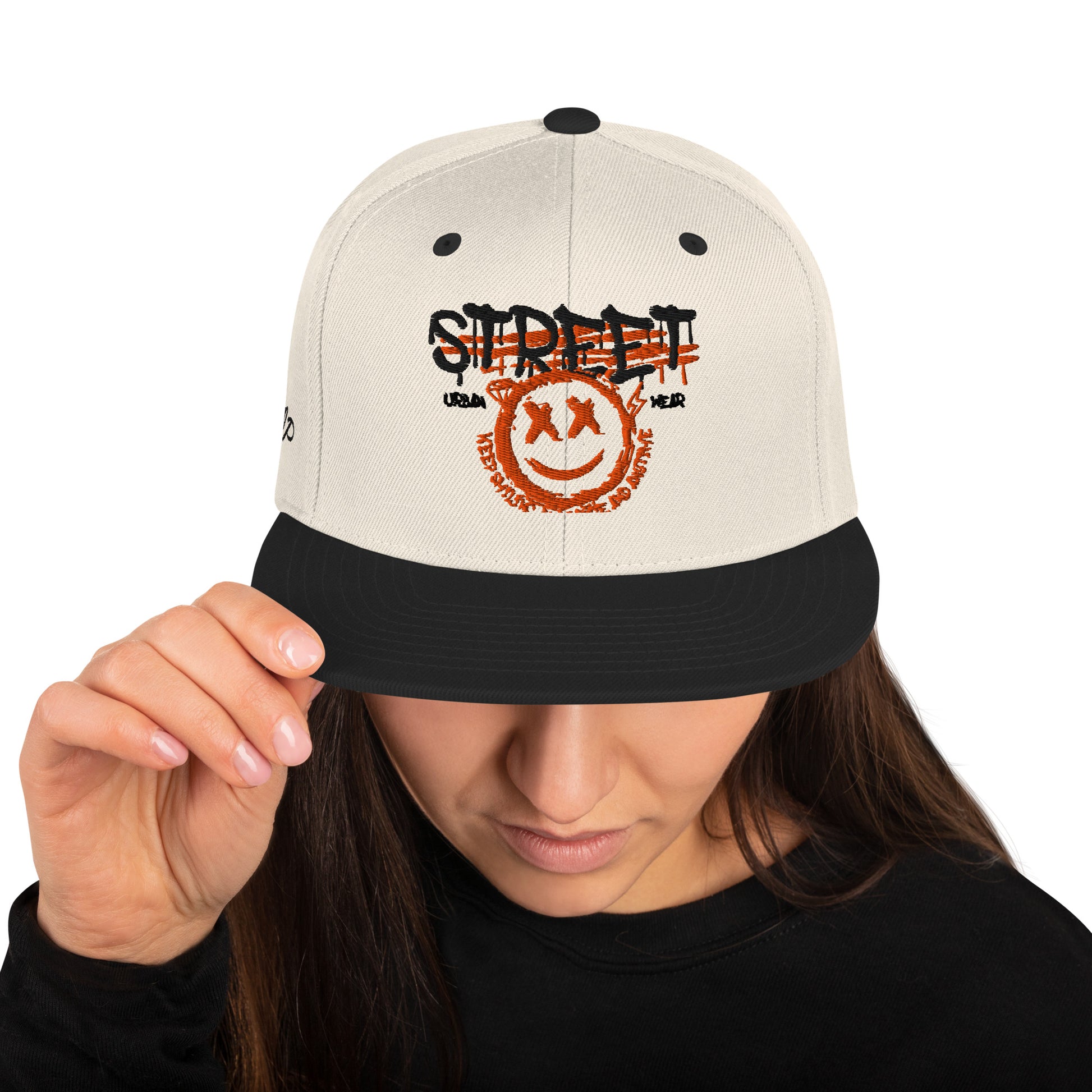 Una donna con i capelli scuri che indossa un cappellino snapback color crema con visiera nera. Il cappellino presenta un design frontale con la scritta 'STREET' in stile graffiato e un'emoji sorridente con occhi a 'X'. Ideale per un look streetwear e skater. Parole chiave: cappellino, cappellino snapback, streetwear, moda skater, accessori moda, cappellino crema e nero, design grafico, abbigliamento urbano, tendenze moda Italia.