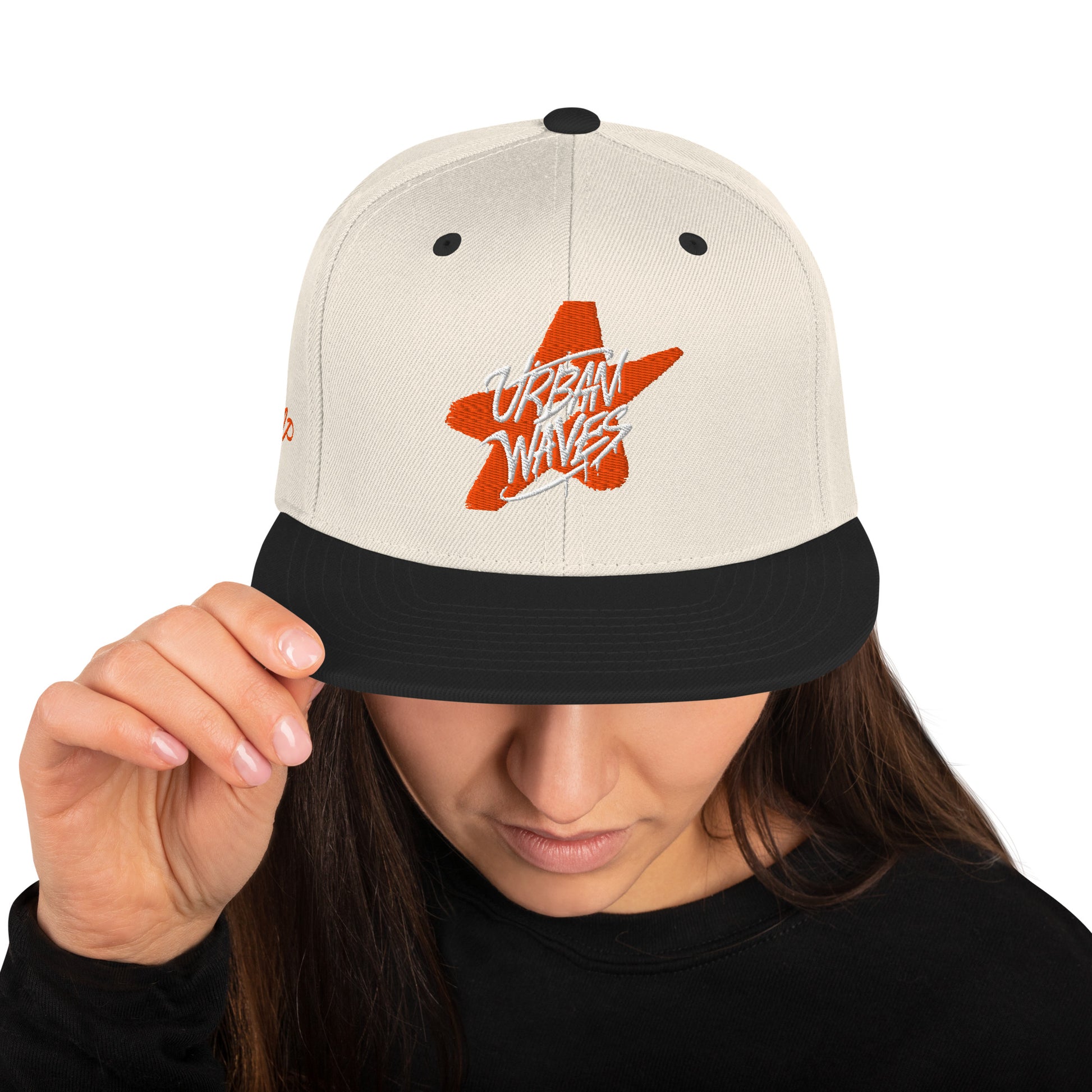 Donna che indossa un cappellino snapback bianco sporco con visiera nera, ricamato con una stella arancione stilizzata e la scritta "URBAN WAVES" in bianco. Un logo aggiuntivo è ricamato sul lato sinistro. Ideale per un look streetwear e casual. Parole chiave: cappellino, snapback, bianco, nero, arancione, stella, urban, streetwear, moda donna, accessori, stile, tendenza, Italia.